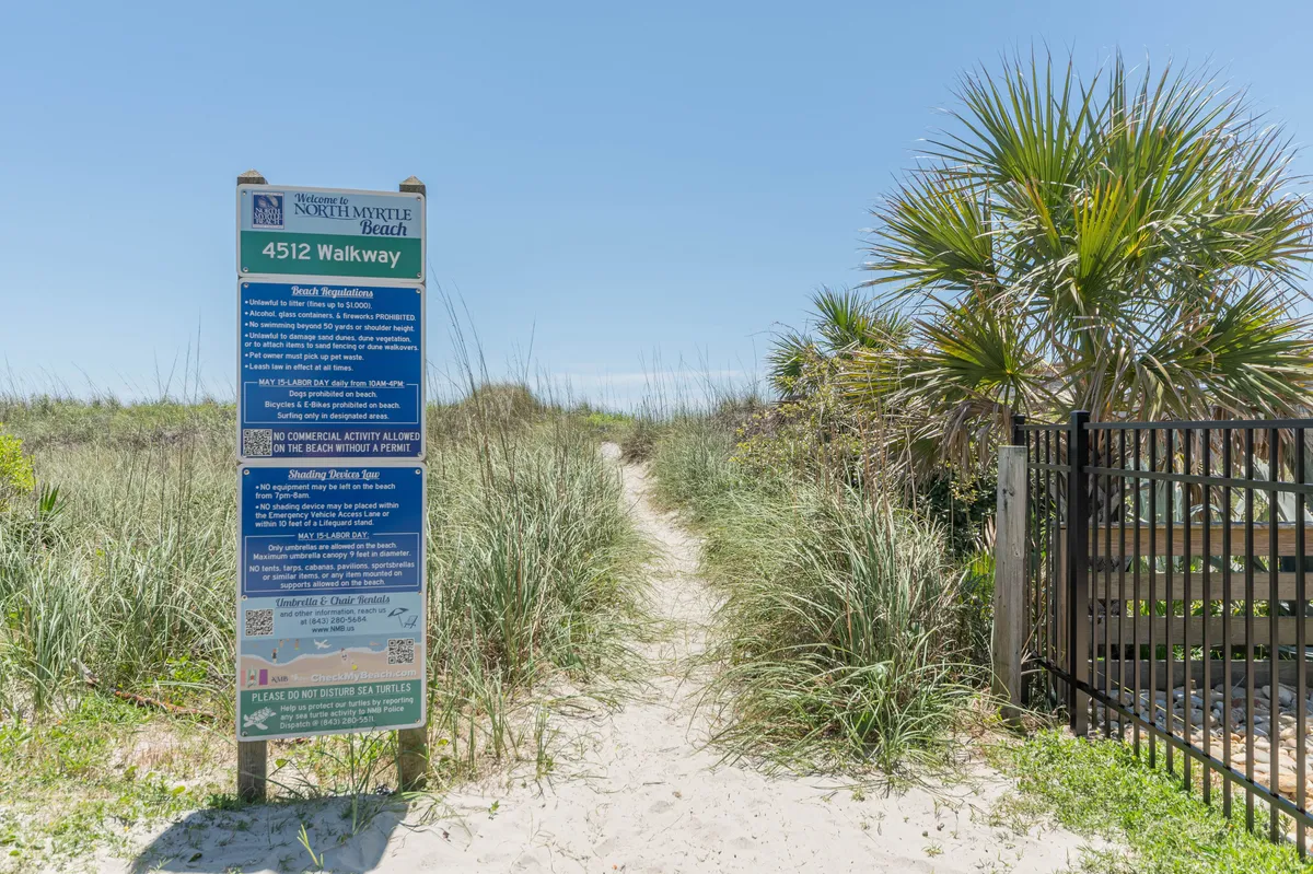 A sign with property information stands beside a pathway lined with greenery, leading toward the beach. The view hints at a tranquil, easy walk from the condo to the sand, reinforcing the narrative of convenience and relaxation.