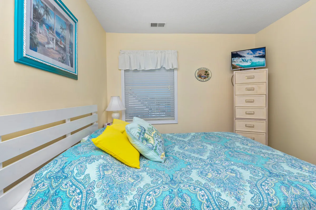 A cozy bedroom with a bed dressed in patterned bedding, a TV placed on a dresser, a dressing table, and framed artwork on the wall. Sunlight gently filters through a window, creating an inviting and peaceful ambiance.