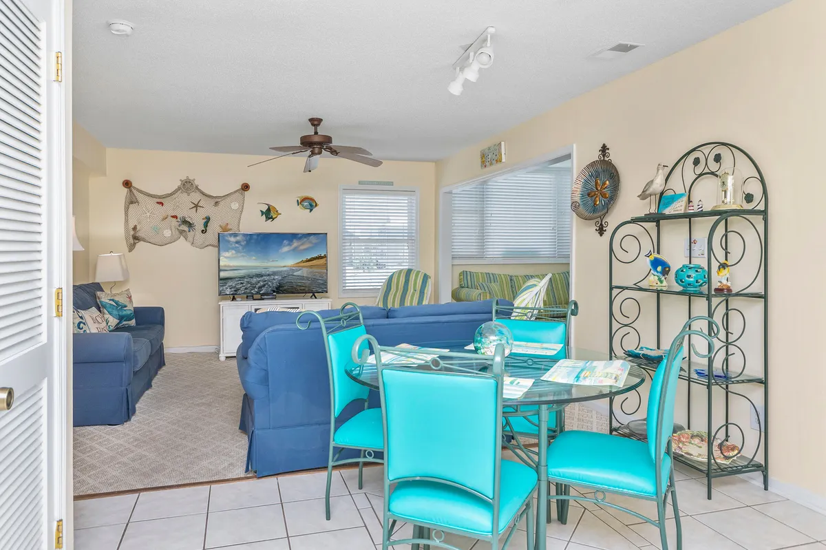 A cheerful living room featuring blue sofas, a glass dining table, decorative wall art, and a television. The space is filled with natural light and arranged for both casual dining and relaxation, inviting guests to gather for movies or meals.