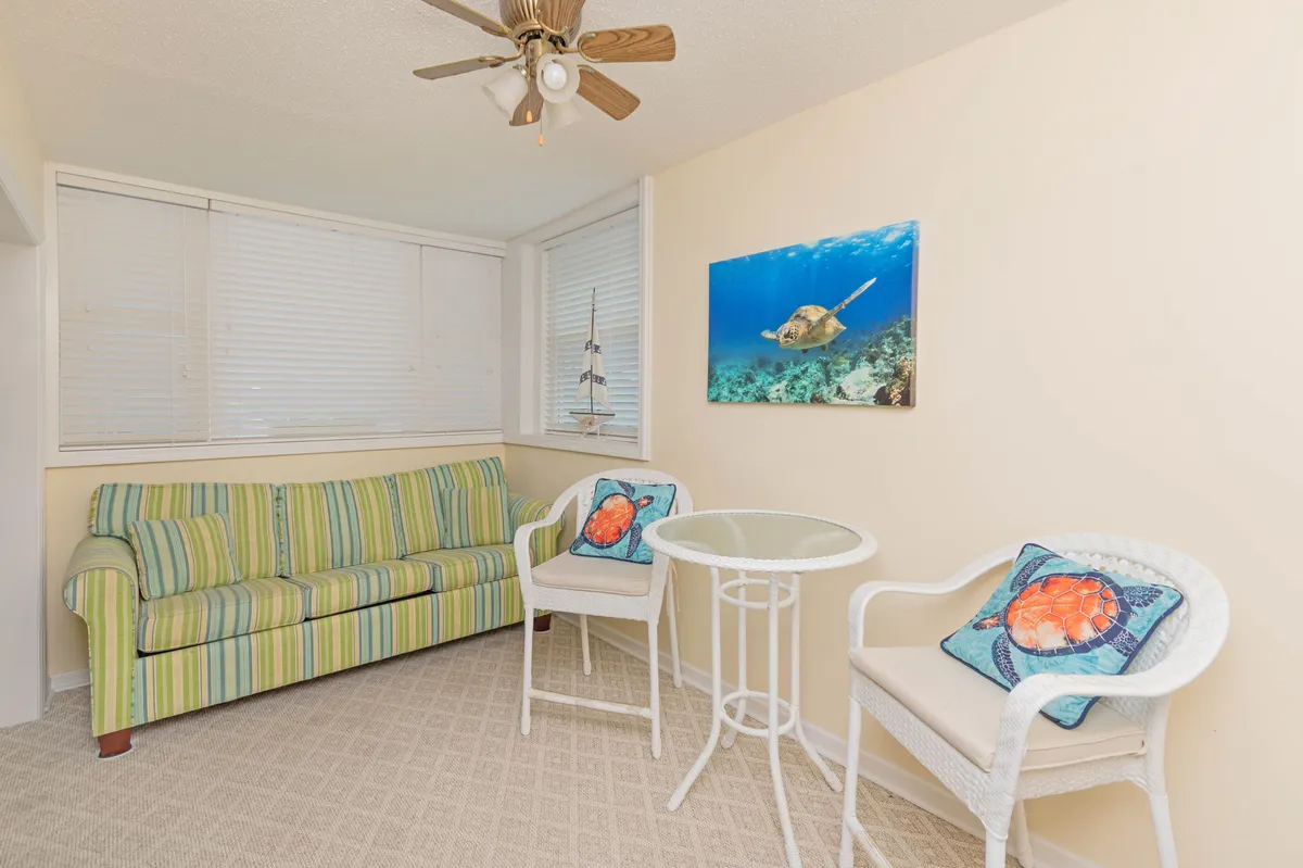 A light-filled sitting area with a striped couch, marine-themed wall art, white wicker chairs, and a glass table. The space exudes a laid-back, coastal vibe, perfect for relaxing or enjoying good company, and hints at a connection with the outdoors through its décor and natural light.