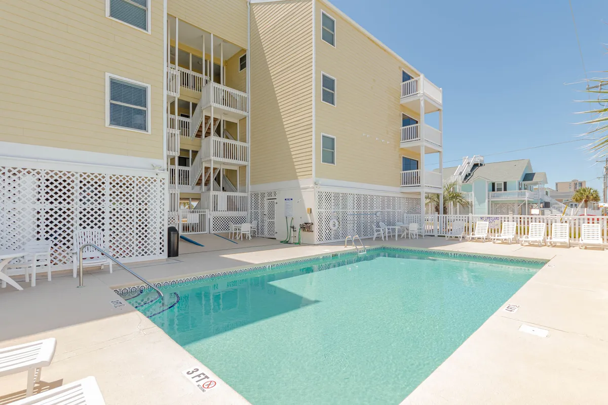 A sparkling swimming pool sits beside the condo building, surrounded by lounge chairs for sunbathing and relaxing after a day at the beach. The inviting pool area is set in the open air, perfect for rinsing off and unwinding outdoors.