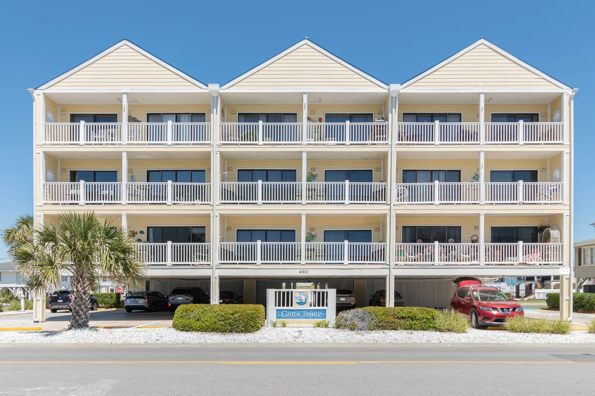 The image shows the exterior of a welcoming three-story condo building, framed by a stately palm tree and an inviting entrance sign. Balconies overlook the entrance, hinting at relaxing mornings and vibrant evenings, while the beach is just a short stroll beyond. The atmosphere is warm and inviting, perfect for guests seeking both relaxation and social connection.