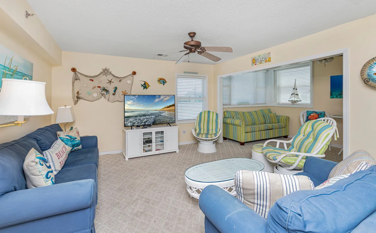 A cozy living area centered around a blue couch decorated with lively pillows, accompanied by colorful striped chairs and a large TV displaying a scenic image. Decorative fish art on the wall adds a playful, beachy vibe, and the space is arranged to facilitate conversation, games, and relaxed group activities.