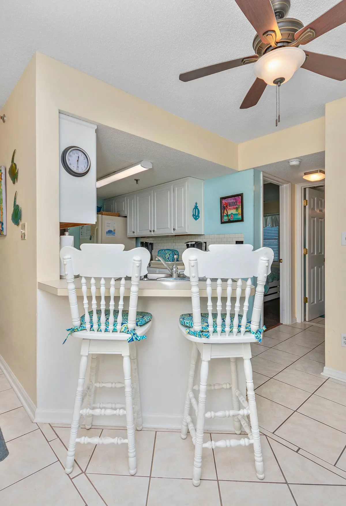 A lively kitchen and dining area with white bar stools at a kitchen counter, vibrant wall art, and a ceiling fan. The setting is perfect for casual meals, socializing, and starting the day with coffee in a bright space.