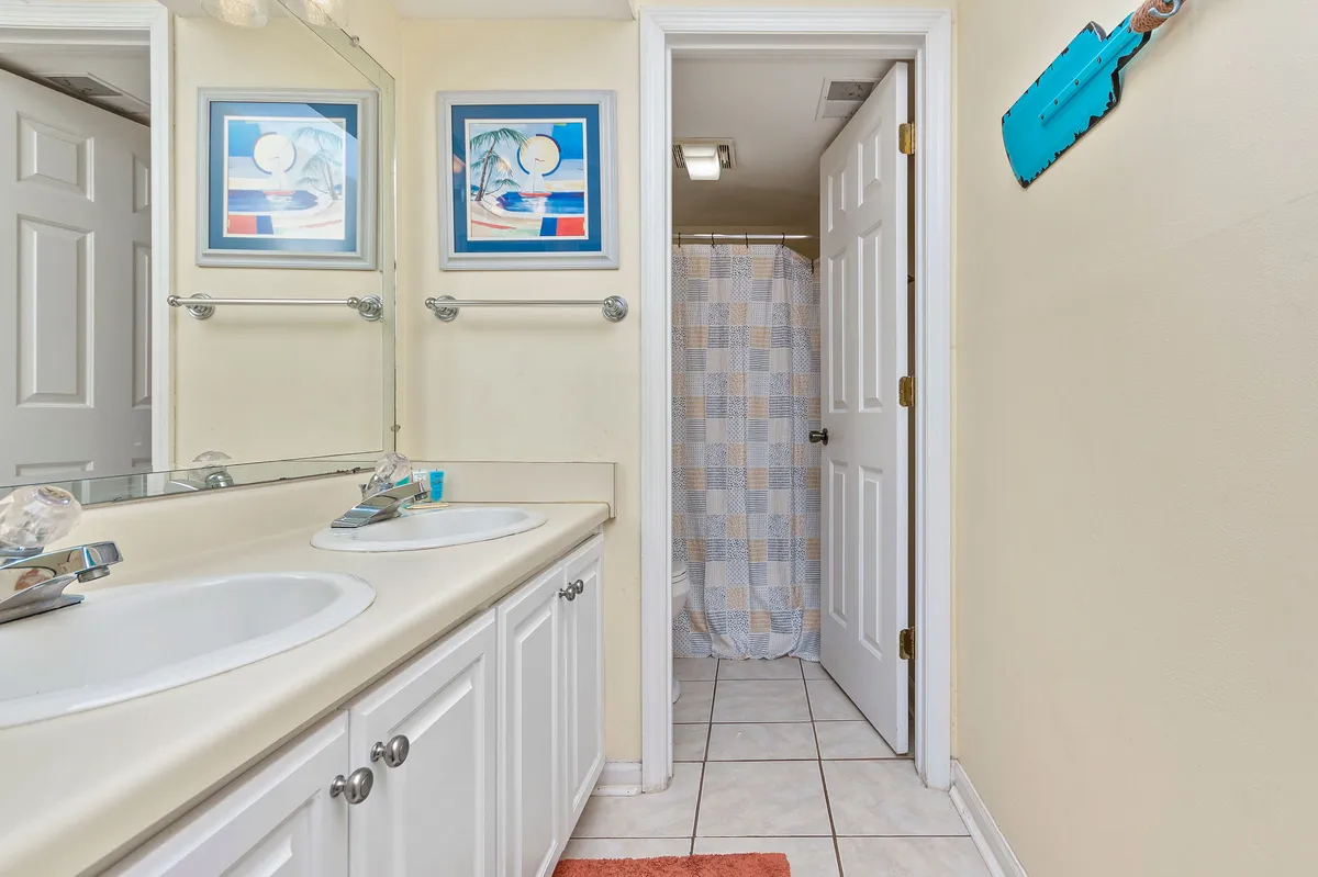 A modern bathroom featuring a double sink with a wide mirror, colorful artwork, and a shower curtain. The space is bright and refreshing, ideal for morning routines or winding down after a day at the beach.