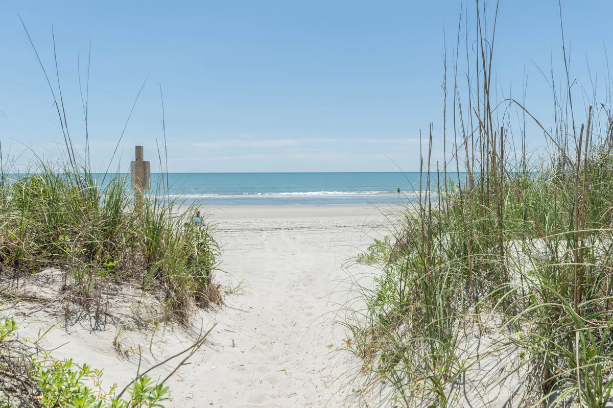 A sandy path lined with dune grass leads directly to the ocean, with waves visible in the distance and a wooden post marking the way. The scene captures the quintessential beach approach, inviting guests to imagine stepping out for sunrise strolls or enjoying a day on the sand.